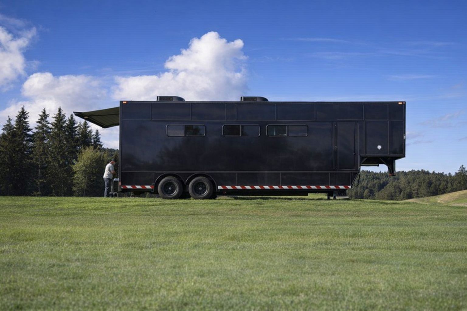 Großer schwarzer Tour-Anhänger auf einer grünen Wiese vor Waldkulisse.